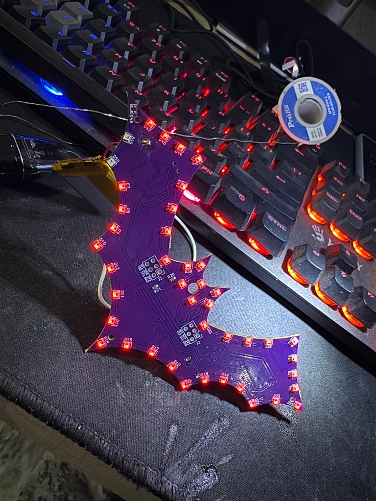 Bat-shaped ESP32 badge PCB with purple solder mask, its perimeter RGB LEDs lit red, running from a connected LiPo cell